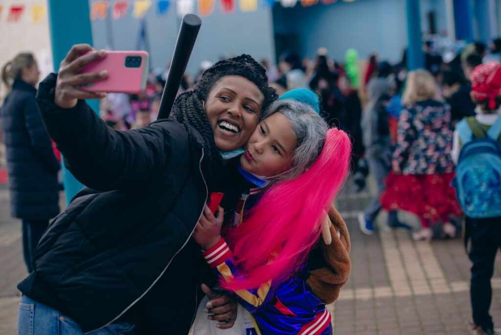 African mother taking a selfie with her multiracial daughter at a school Purim celebration. Pupils celebrate Purim carnival with costumes. Festival of Purim.